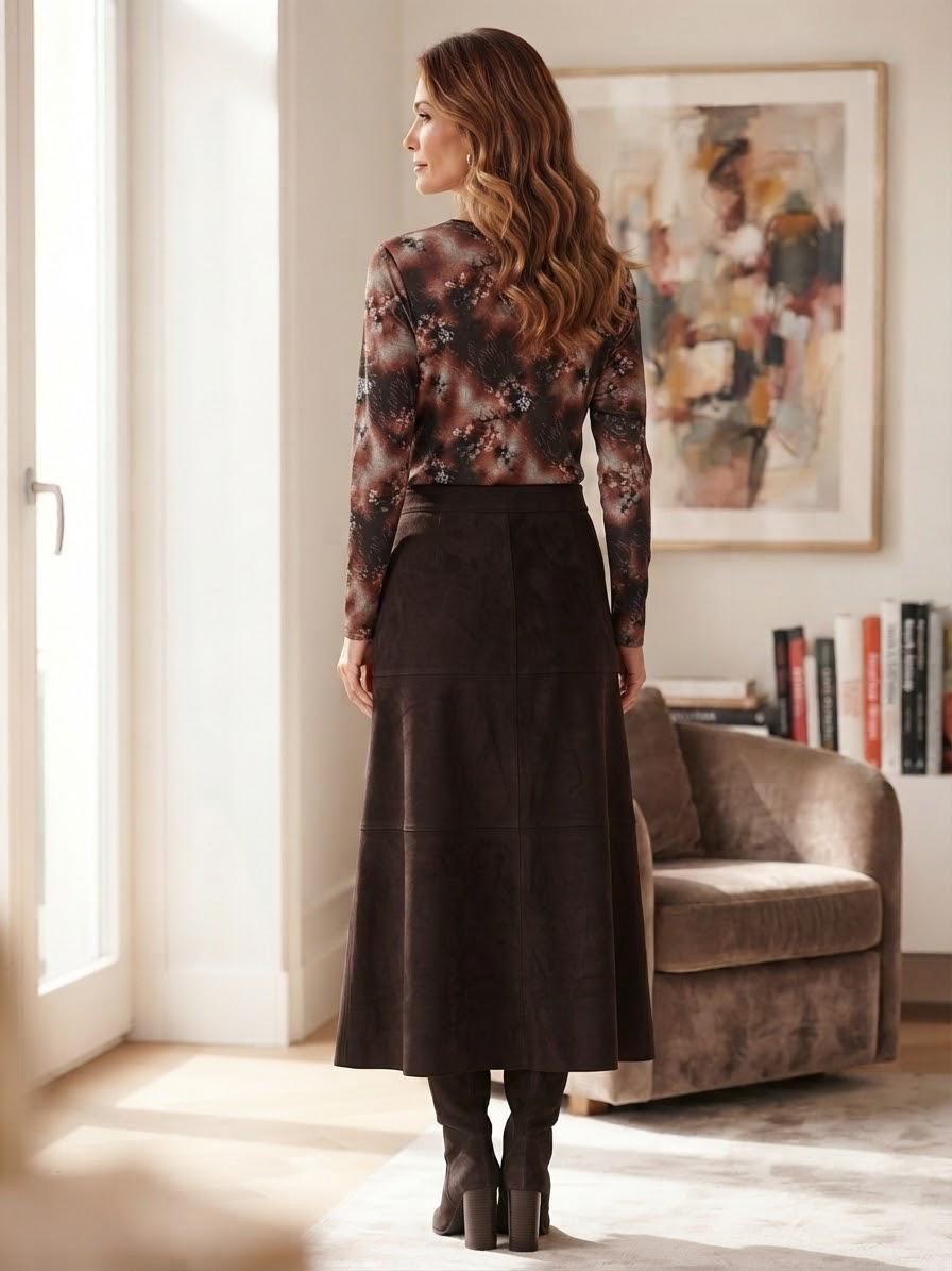 A woman stands in a sunlit room wearing a long-sleeve floral top and a dark brown midi skirt, shown from the back. A brown armchair, framed abstract wall art, and books are visible in the background.
