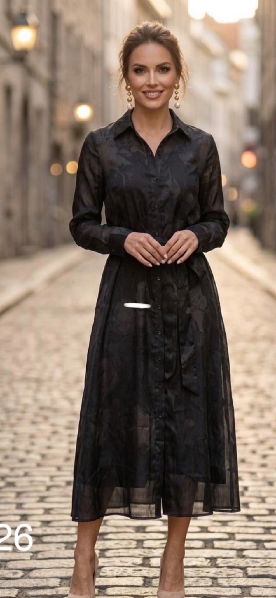 A woman modeling a black sheer long-sleeve midi shirt dress with a subtle tonal pattern, standing on a cobblestone street.